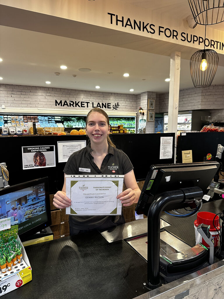 Gemma standing behind a cash register, proudly holding a certificate and smiling.