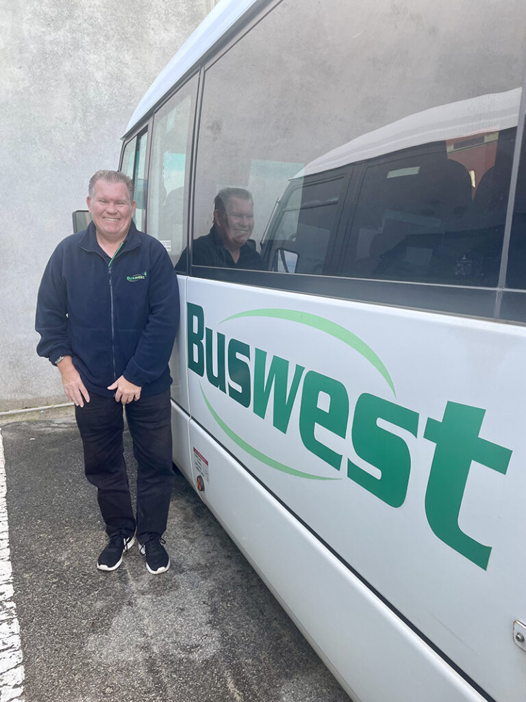 Mark standing alongside a white bus branded with the Buswest logo."