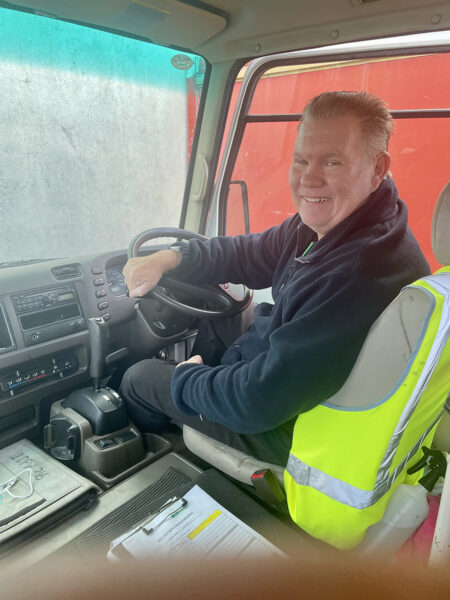 Mark sitting in the drivers seat of the bus with one hand on the steering. He is looking over his shoulder and smiling."