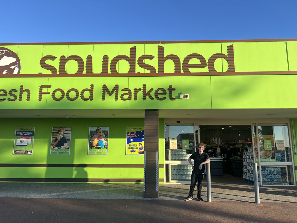 Sean standing in front of the Spudshed stop front of a green wall and the words, 'Spudshed. Food market.'