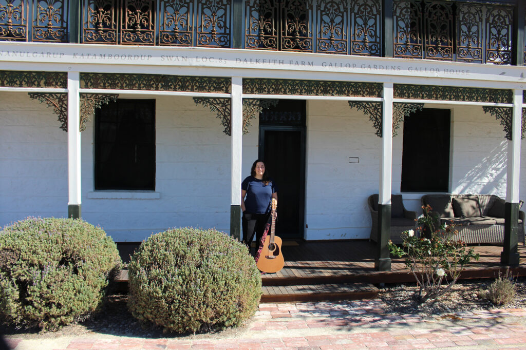 A woman standing on a porch with her guitar in front of her.