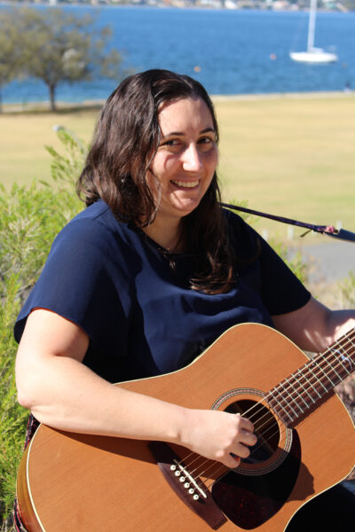 alt="Suzanne sitting down holding her guitar in front of a lovely background of grass and water."