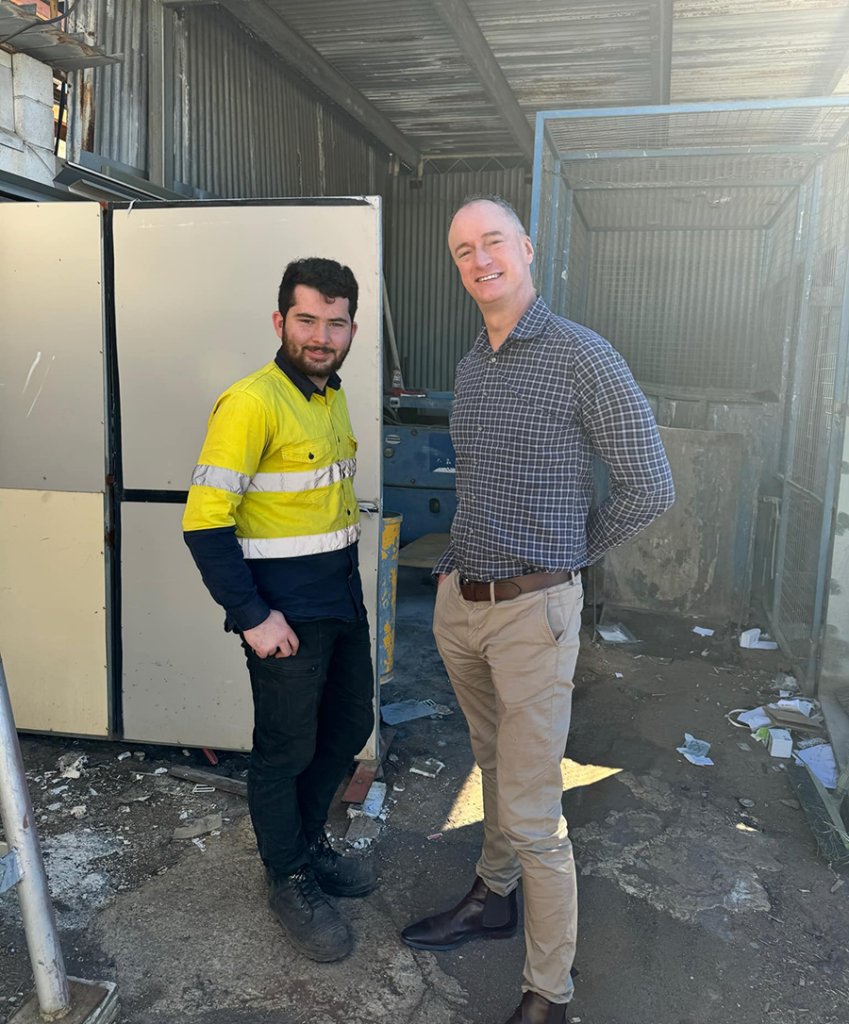 Two men standing next to each other in a shed. Joel on the left is wearing high-vis and Dan on the right is dressed smart-casual.