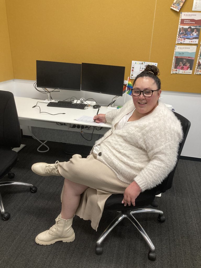 Chloe sitting at a desk while holding a pen to a piece of paper and smiling.