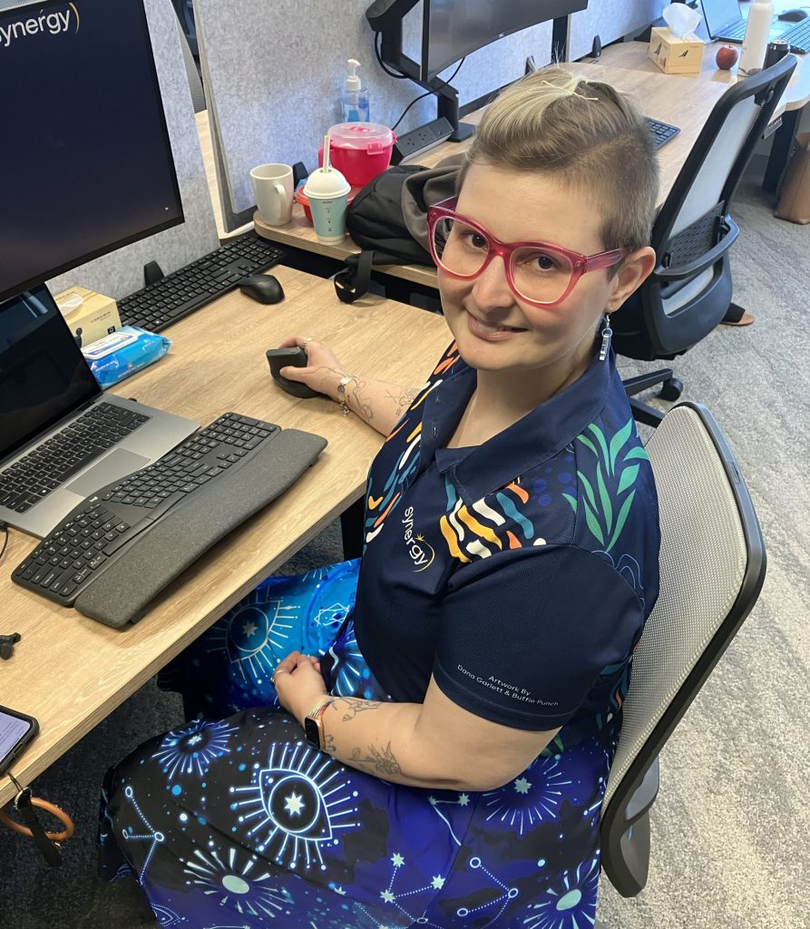 Eloise sitting at a desk, wearing pink glasses and smiling at the camera.