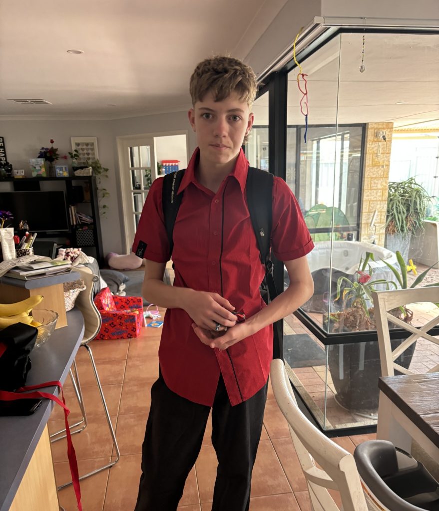 A young boy wearing a backpack and smiling in a red button up t-shirt.