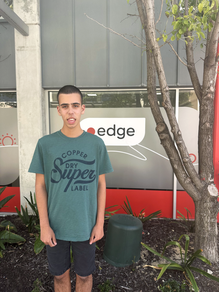 Dermarco is wearing a green shirt while standing in front of the Edge branded windows smiling.