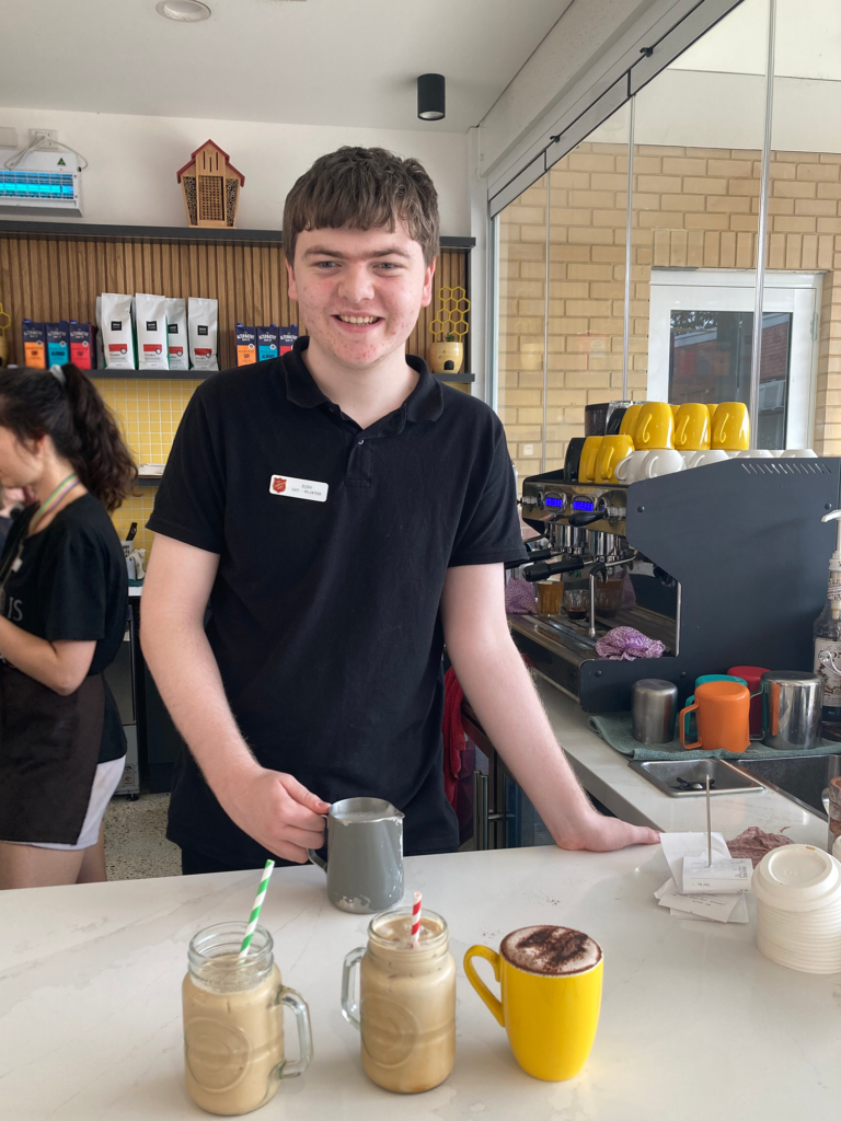 Rory standing behind a the cafe counter with three cups of coffees in front of him, while he holds a metal milk jug.