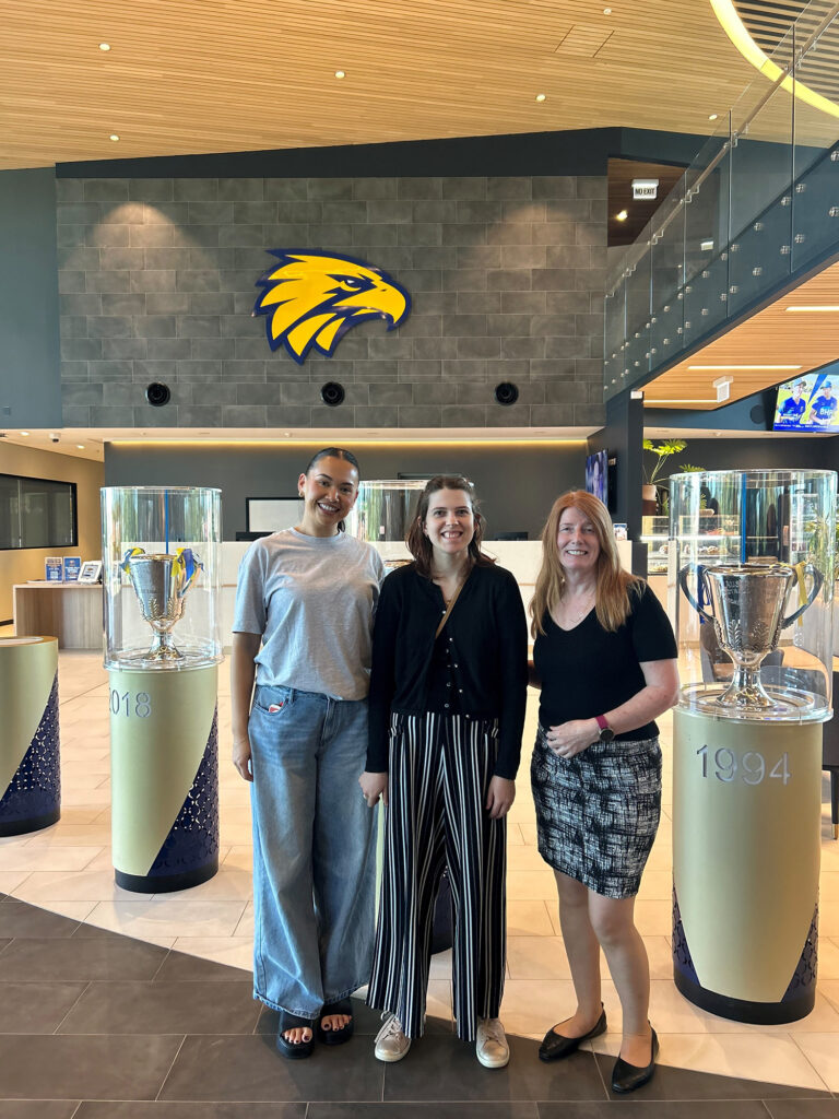 Two women and a man standing in front of a lobby with trophies behind them.