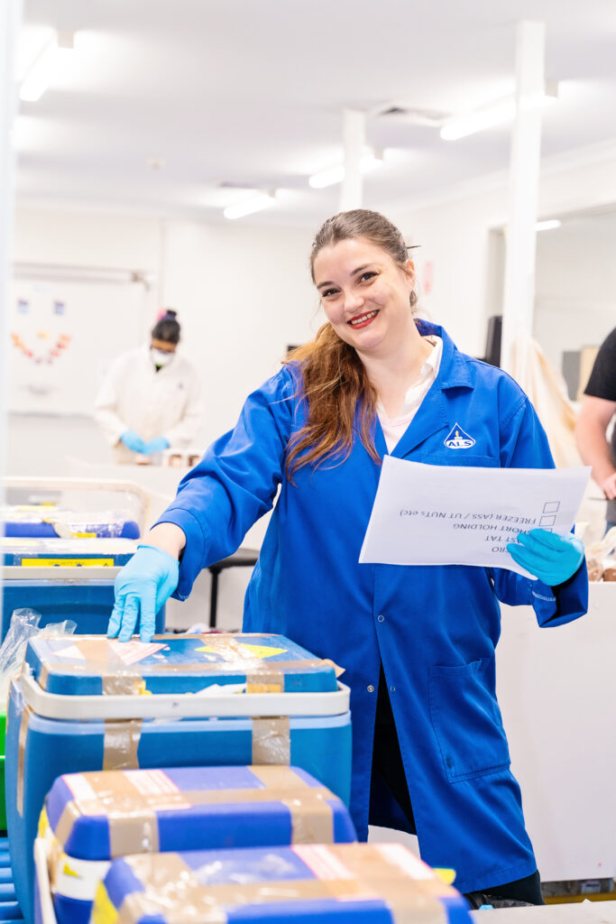 Stephanie holding a piece of paper in one hand while her other hand is resting on a blue container. She is smiling for a photo in her urse in blue scrubs holding a piece of paper and donating boxes of supplies.